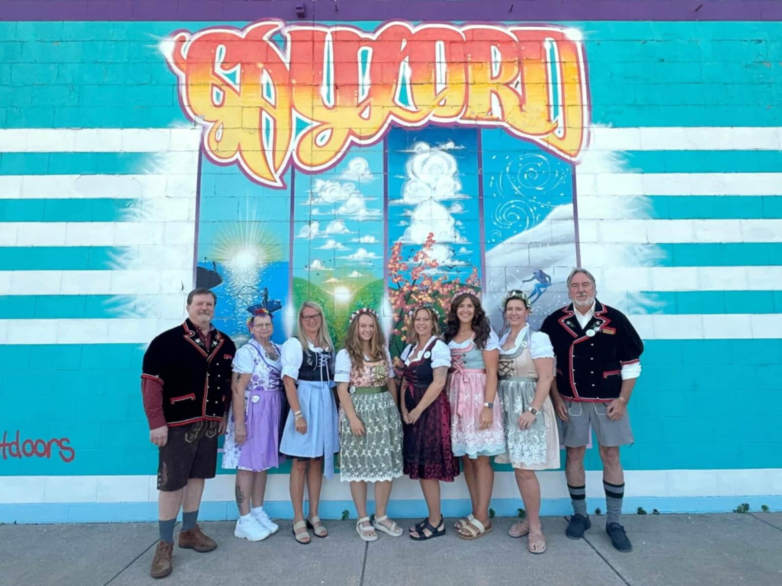 Gaylord Tourism Bureau & Chamber of Commerce Staff wearing lederhosen and dirndls for Alpenfest in front of the "Gaylord" mural painted onto a downtown building.